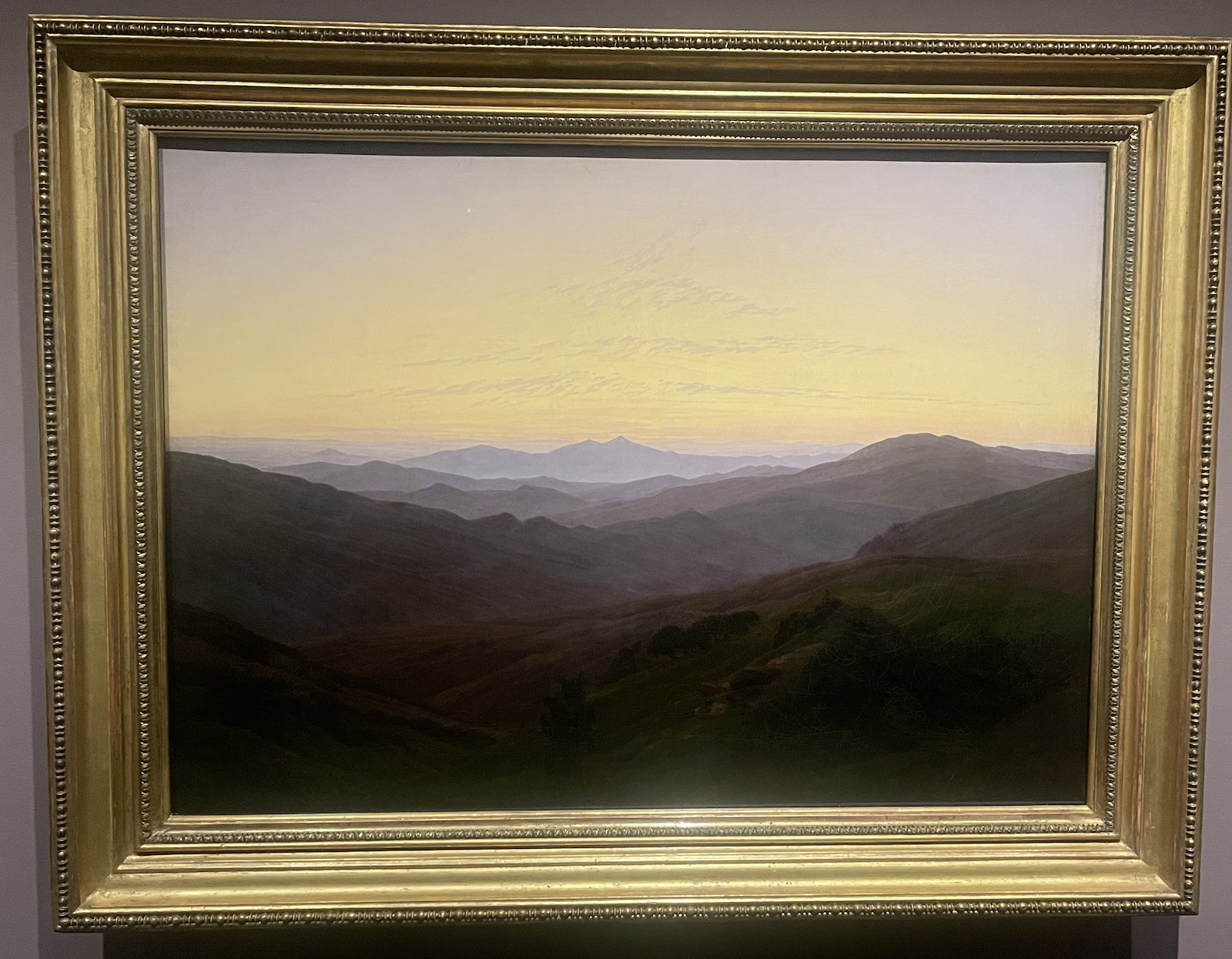 Friedrich distilled the topography of the Giant Mountains to undulating waves of green, brown, and gray that recede rhythmically into the distance. The muted earth tones provide a foil for the nuanced tints of the sky. Although at first glance the landscape appears to be uninhabited, a closer look shows a man seated on a rock to the right of the trees—a tiny figure almost lost within the boundless landscape.